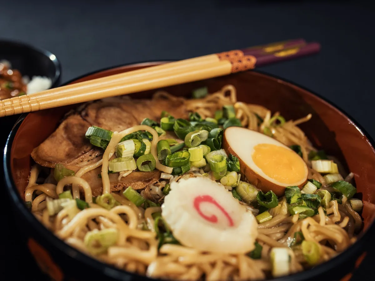 Nahaufnahme einer Schale Ramen mit Chashu-Schweinefleisch, einem halben weich gekochten Ei, einem rosa geringelten Fischkuchen und gehackter Frühlingszwiebel unter Holzstäbchen