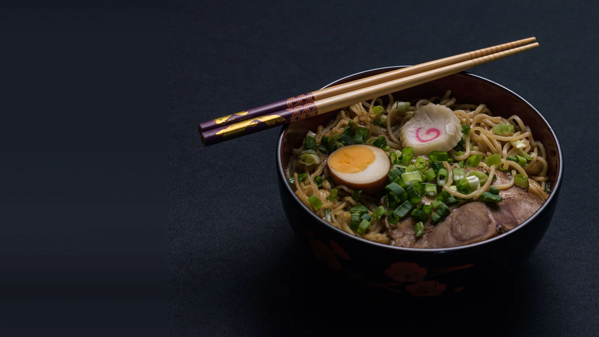 Eine schwarze Lackschale mit Ramen, dazu ein halbiertes weich gekochtes Ei, ein rosa geringeltes Fischkuchenstück, gehackte Frühlingszwiebeln und geschnittenes Schweinefleisch, Holzstäbchen liegen über dem Rand, auf einem dunklen Tuch